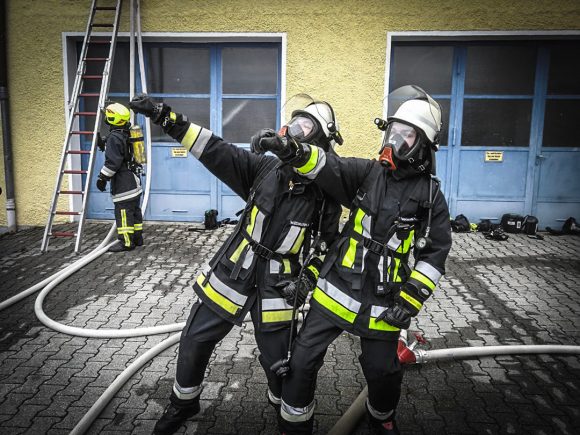 Zwei Feuerwehrleute in Schutzkleidung zeigen mit Schläuchen in einer Feuerwehrstation.