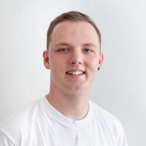 Junger Mann mit kurzem Haar und einem weißen T-Shirt, der in die Kamera lächelt.