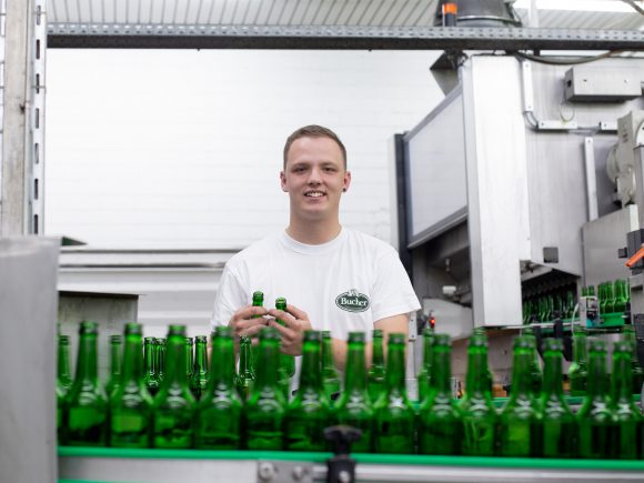 Mitarbeiter in einem Brauerei mit grünen Flaschen auf einem Förderband, lächelnd in einem weißen T-Shirt.