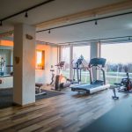 Fitnessraum mit Laufband, Crosstrainer und weiteren Trainingsgeräten vor großen Fenstern.