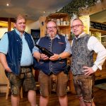Drei Männer in traditioneller bayerischer Tracht halten eine Weinflasche in einem Innenraum.