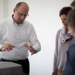 Ein Mann in einem weißen Hemd erklärt etwas vor einer Gruppe von Personen in einem hellen Raum.