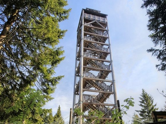 Holzturm mit mehreren Etagen und Wendeltreppe, umgeben von Bäumen im Wald.