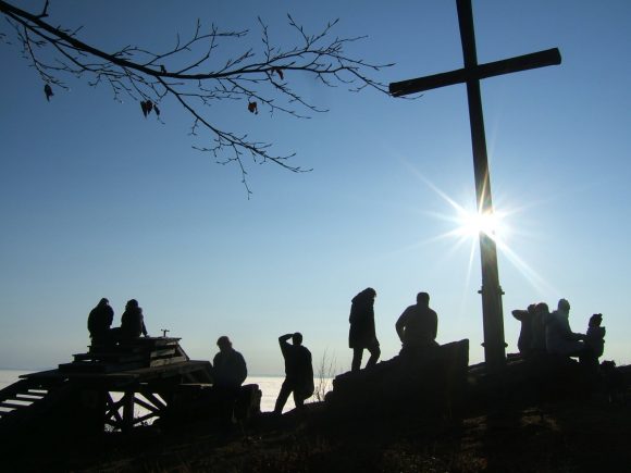 Silhouetten von Menschen, die vor einem großen Kreuz auf einem Hügel stehen, mit Sonnenstrahlen im Hintergrund.