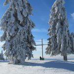 Zwei große, schneebedeckte Bäume mit einem Holzkreuz im Hintergrund auf einem Berg.