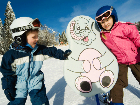 Zwei Kinder in Skiausrüstung stehen neben einem Skischul-Schild mit einem Elefantenmotiv.
