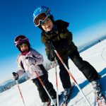 Zwei Kinder in Skiausrüstung auf einer Skipiste im Winter mit blauen Himmel.