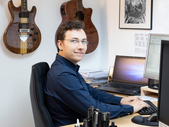 Mann mit Brille sitzt an einem Schreibtisch mit Computer und Gitarren im Hintergrund.
