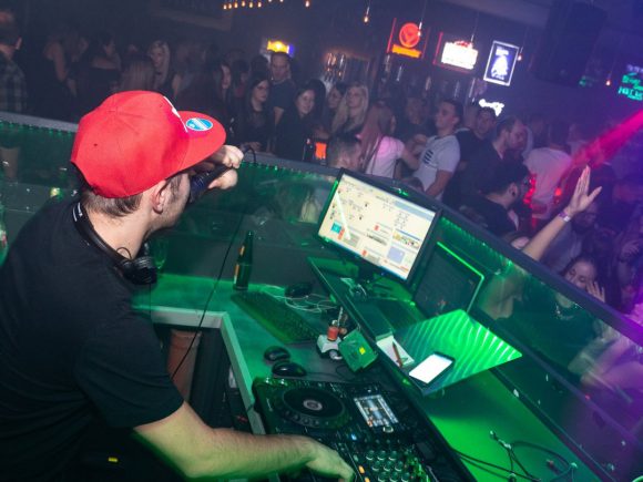 DJ mit roter Mütze, der an einem Mischpult in einem Club auflegt, während das Publikum tanzt.