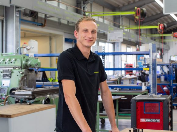 Junger Mann mit kurzem Haar in einem schwarzen Poloshirt steht an einem Tisch in einer Werkstatt.