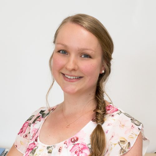 Frau mit langen, geflochtenen Haaren und einer Bluse mit Blumenmuster lächelt in die Kamera.