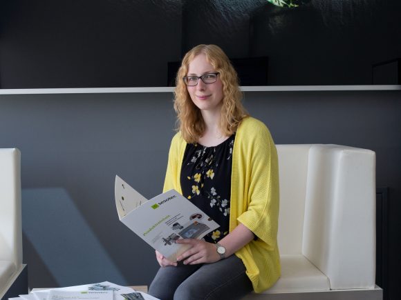 Eine Frau mit blonden Haaren und Brille sitzt auf einem weißen Stuhl und hält eine Broschüre in der Hand.