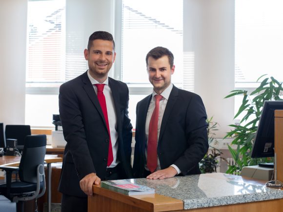 Zwei Männer in Anzügen stehen am Empfangstresen eines Büros mit Computer und Pflanzen im Hintergrund.