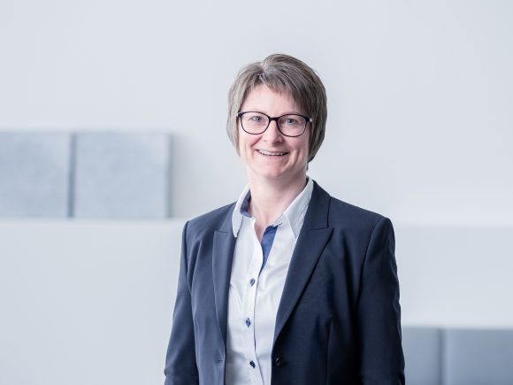 Frau mit kurzen Haaren und Brille trägt einen dunklen Anzug und steht vor einem hellen Hintergrund.