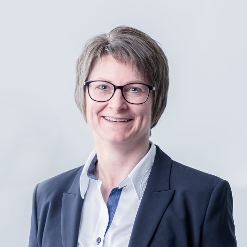 Frau mit kurzen, grauen Haaren und Brille, die in einem Anzug lächelt.