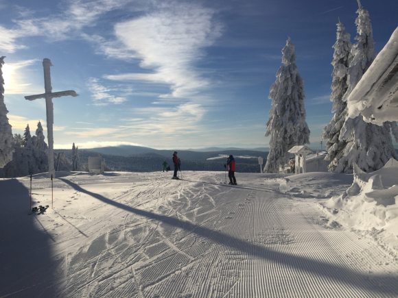 Zwei Skifahrer auf einer verschneiten Piste mit Bäumen und einem Kreuz im Hintergrund.