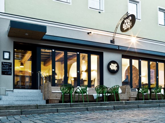 Fassade eines Restaurants mit großen Fenstern und grünen Stühlen im Außenbereich.
