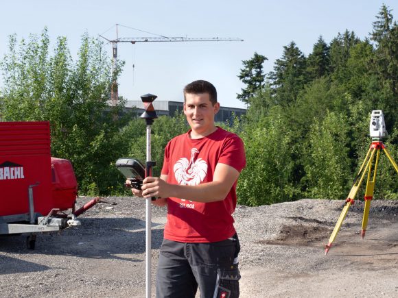 Junger Mann in rotem T-Shirt hält ein Vermessungsgerät auf einer Baustelle mit Bäumen im Hintergrund.