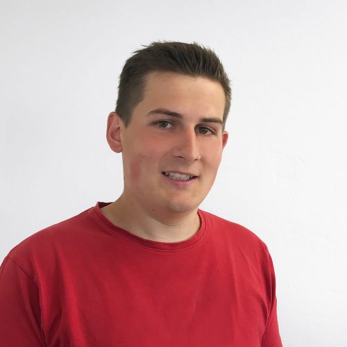 Ein junger Mann mit kurzen Haaren trägt ein rotes T-Shirt und lächelt in die Kamera.