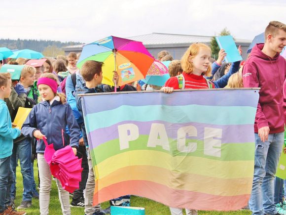 Schüler halten eine Regenbogenflagge mit dem Wort 'PACE' während einer Demonstration im Freien.