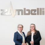 Zwei Frauen stehen nebeneinander vor einem Zambelli-Schild in einem Innenraum.