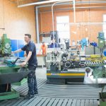 Ein Mitarbeiter bedient eine Fräsmaschine in einer Maschinenbauwerkstatt mit weiteren Personen im Hintergrund.
