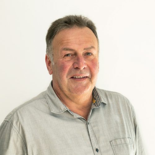 Ein Mann mit grauem Hemd und lächelndem Gesicht vor einem hellen Hintergrund.
