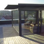Moderne Veranda mit großen Glasfenstern und Holzterrasse, die einen Blick auf die Umgebung bietet.