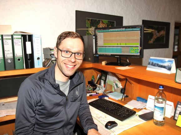 Mann mit Brille sitzt an einem Schreibtisch mit einem Computerbildschirm und Unterlagen.