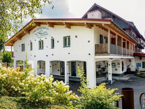 Ein Hotelgebäude mit weißer Fassade, Balkon und Eingangsbereich, umgeben von Pflanzen.