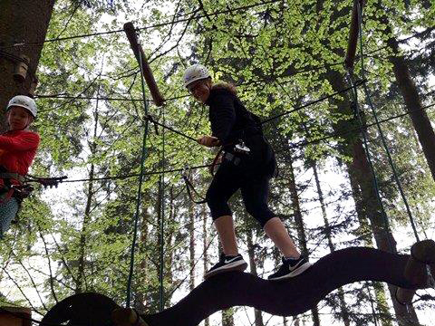 Eine Person balanciert auf einem Kletterelement in einem Wald mit grünen Blättern.