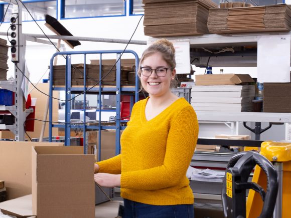 Frau mit Brille und gelbem Pullover, die in einer Verpackungsumgebung an einem Karton arbeitet.