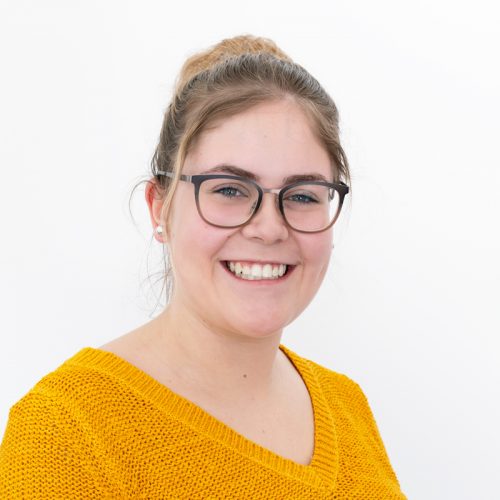 Eine junge Frau mit Brille und einem gelben Pullover lächelt in die Kamera.
