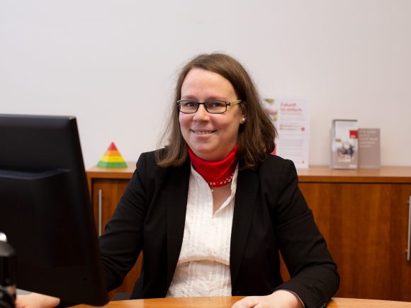 Frau mit Brille und rotem Schal sitzt am Schreibtisch vor einem Computerbildschirm.