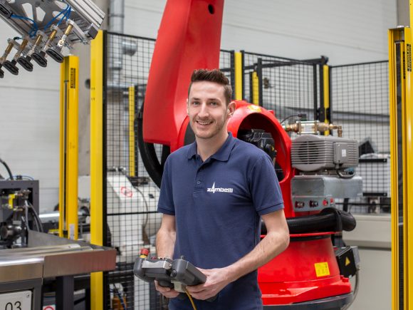 Ein Mitarbeiter hält eine Fernbedienung in einer industriellen Umgebung mit einem Roboter im Hintergrund.