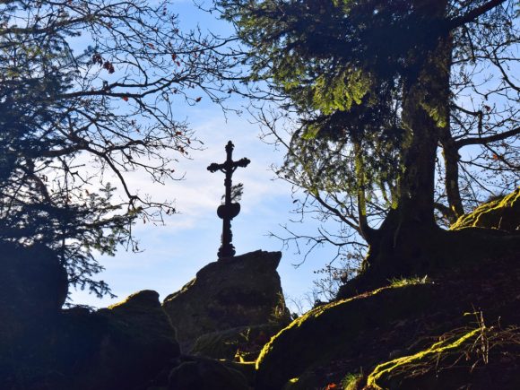 Silhouette eines Kreuzes auf einem Felsen, umgeben von Bäumen und Sträuchern im Wald.