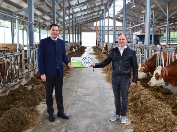 Zwei Männer stehen in einem Stall und halten ein Schild mit dem Logo der Landwirtschaft.