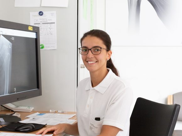 Frau mit Brille sitzt an einem Schreibtisch und betrachtet ein Röntgenbild auf dem Computerbildschirm.