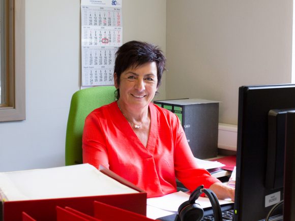 Frau in rotem Oberteil sitzt an einem Schreibtisch mit Computer und Aktenordnern im Hintergrund.