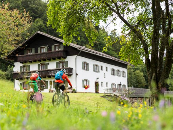 Zwei Radfahrer fahren auf einem Weg, der an einem weißen Haus mit Balkon vorbeiführt.