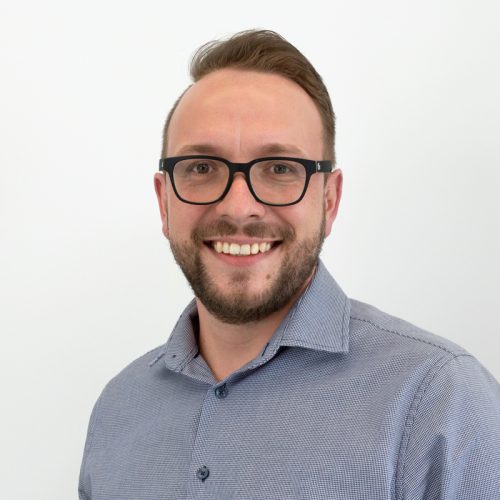 Mann mit Brille und kurzem Bart, der in einem hellen Hemd lächelt und in die Kamera schaut.