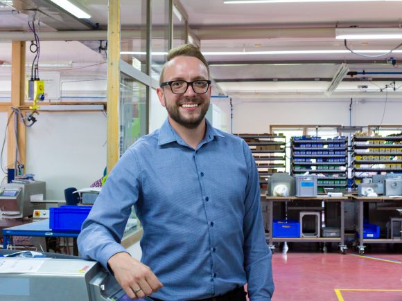 Ein Mann steht in einem modernen Büro mit einem Metall-Detektor im Vordergrund und Regalen im Hintergrund.