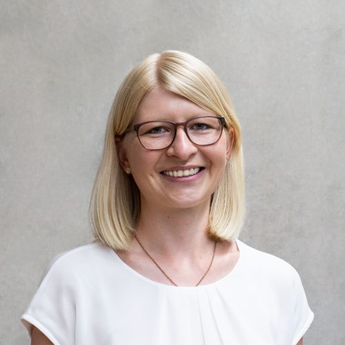 Frau mit blonden Haaren und Brille lächelt vor einer neutralen Wand.