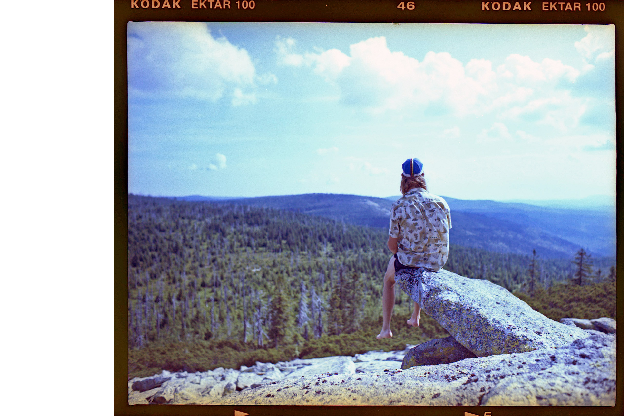 Eine Person sitzt auf einem Felsen und schaut auf eine bewaldete Landschaft unter einem blauen Himmel.