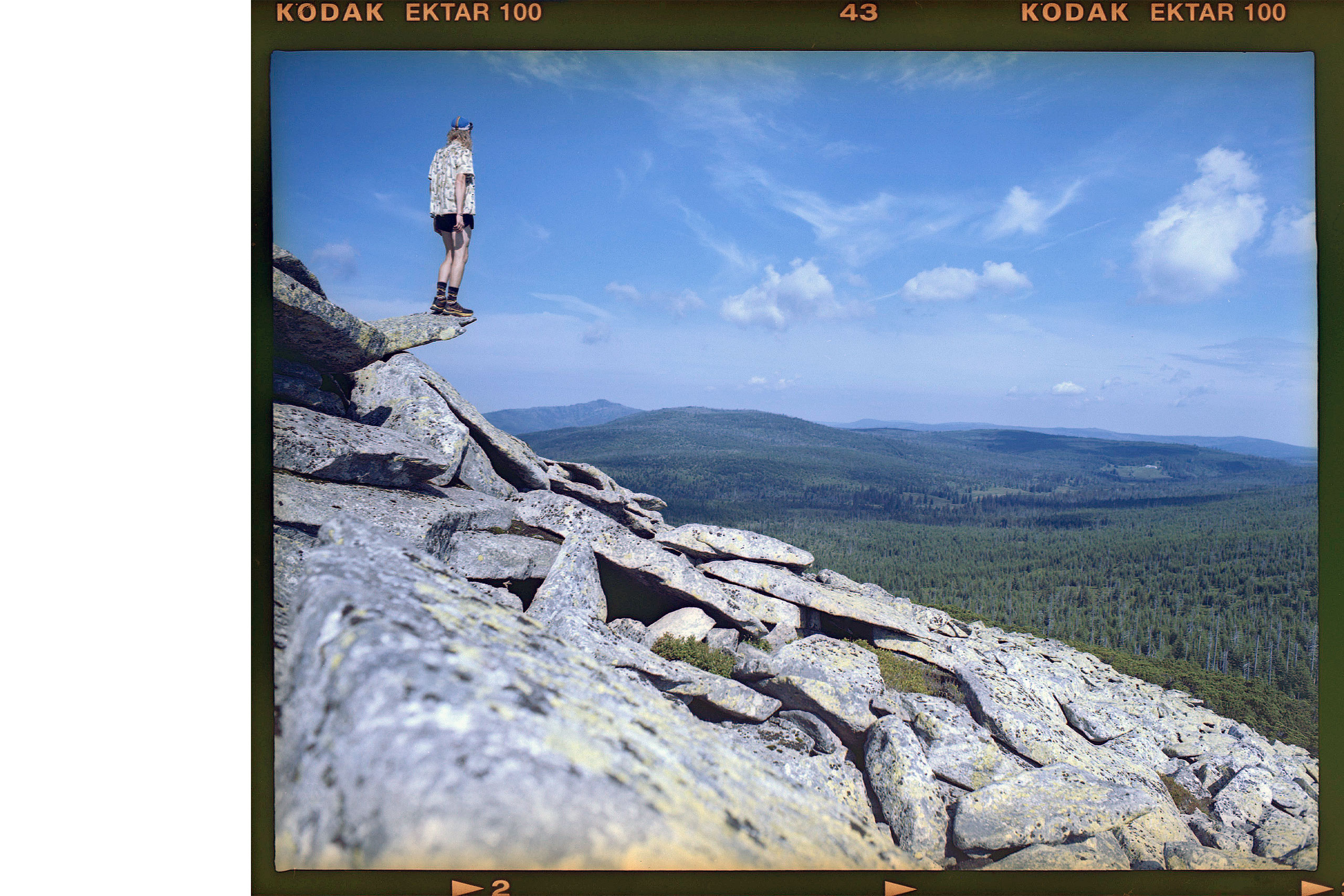 Eine Person steht auf einem Felsen und blickt auf eine weite, bewaldete Landschaft unter einem blauen Himmel.