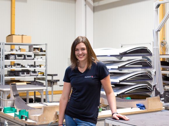 Frau steht in einer Werkstatt neben Bauteilen und Werkzeugen auf einem Tisch.