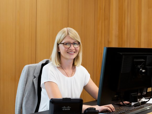 Eine Frau mit blonden Haaren sitzt an einem Schreibtisch und arbeitet an einem Computer.