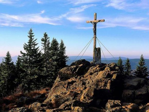 Holzkreuz auf einem großen Felsen, umgeben von Nadelbäumen und einem klaren Himmel.