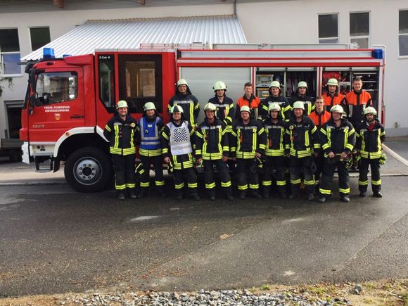 Gruppe von Feuerwehrleuten in Uniform vor einem roten Feuerwehrfahrzeug mit Blaulicht.