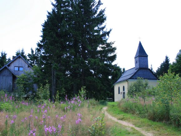 Eine kleine Kirche mit einem spitzen Turm neben einem Holzgebäude in einer bewaldeten Umgebung.